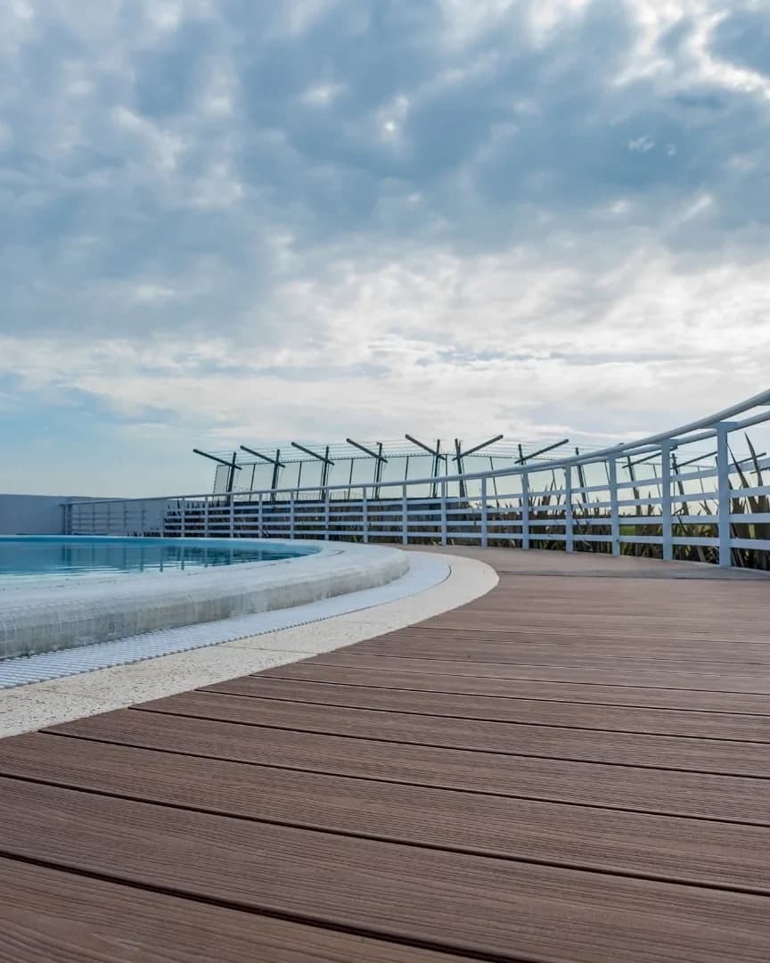 Deck Sólido de WPC café que rodea en curva una alberca de azotea, con barandal blanco y cielo nublado
