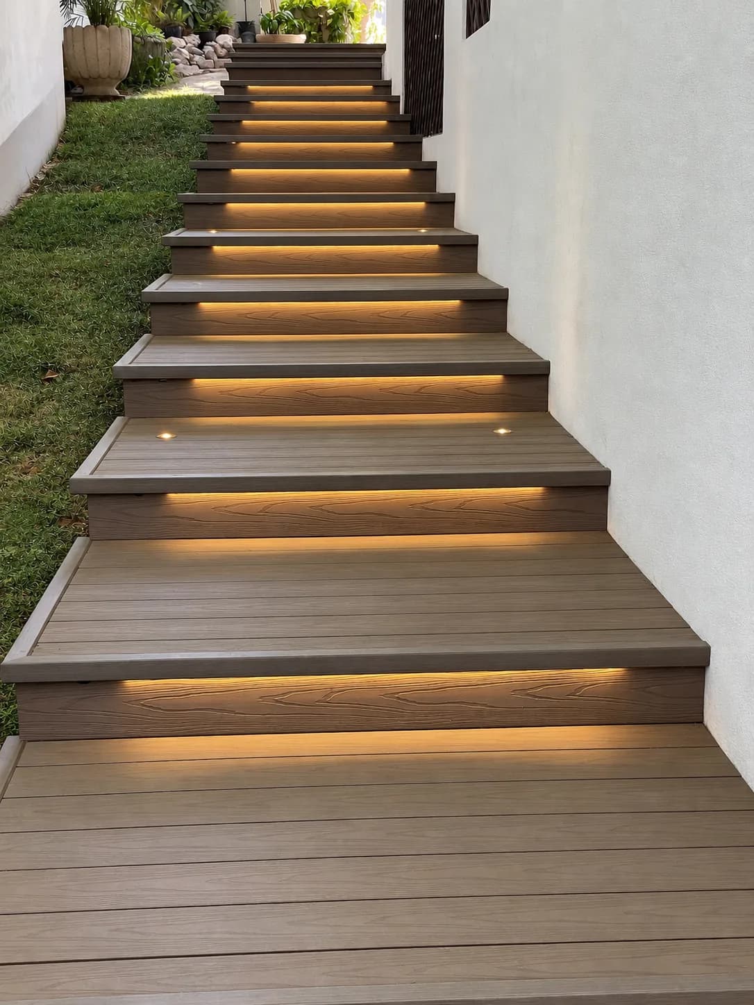 Escalera exterior de deck WPC tono madera con luces empotradas bajo cada escalón, junto a un muro blanco y un talud de pasto