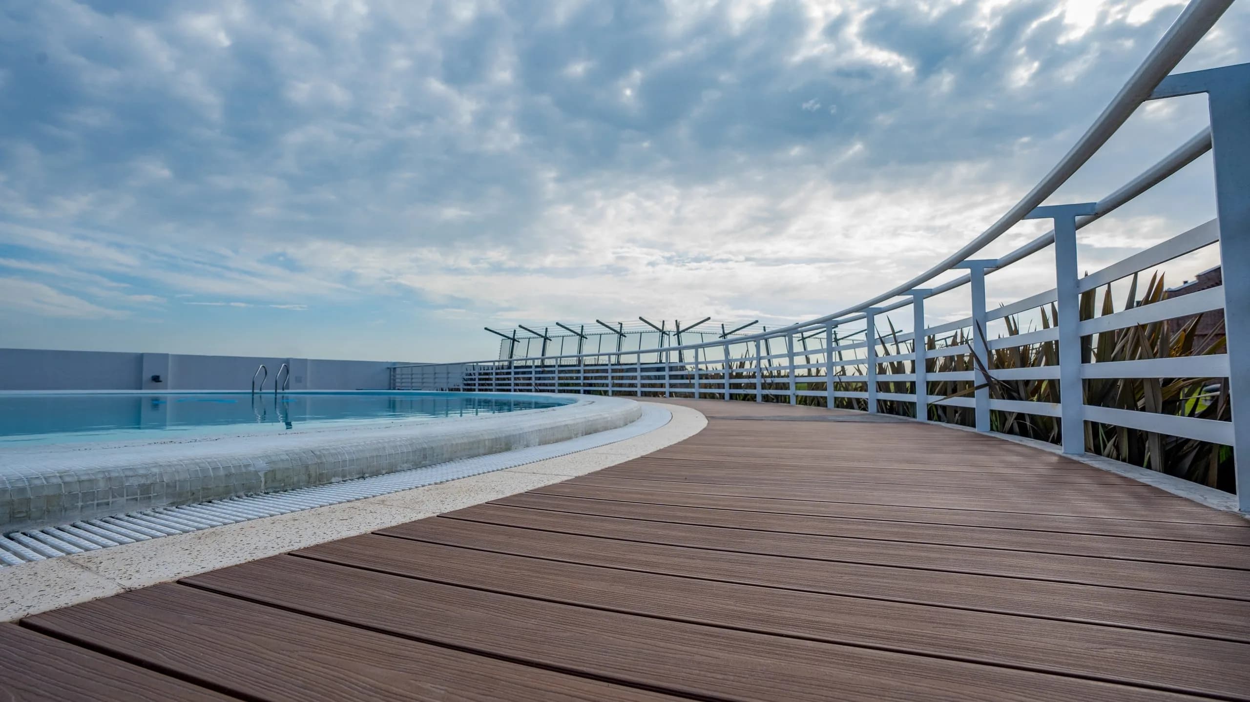 Rooftop with curved WPC decking, a white railing and a pool under a cloudy sky