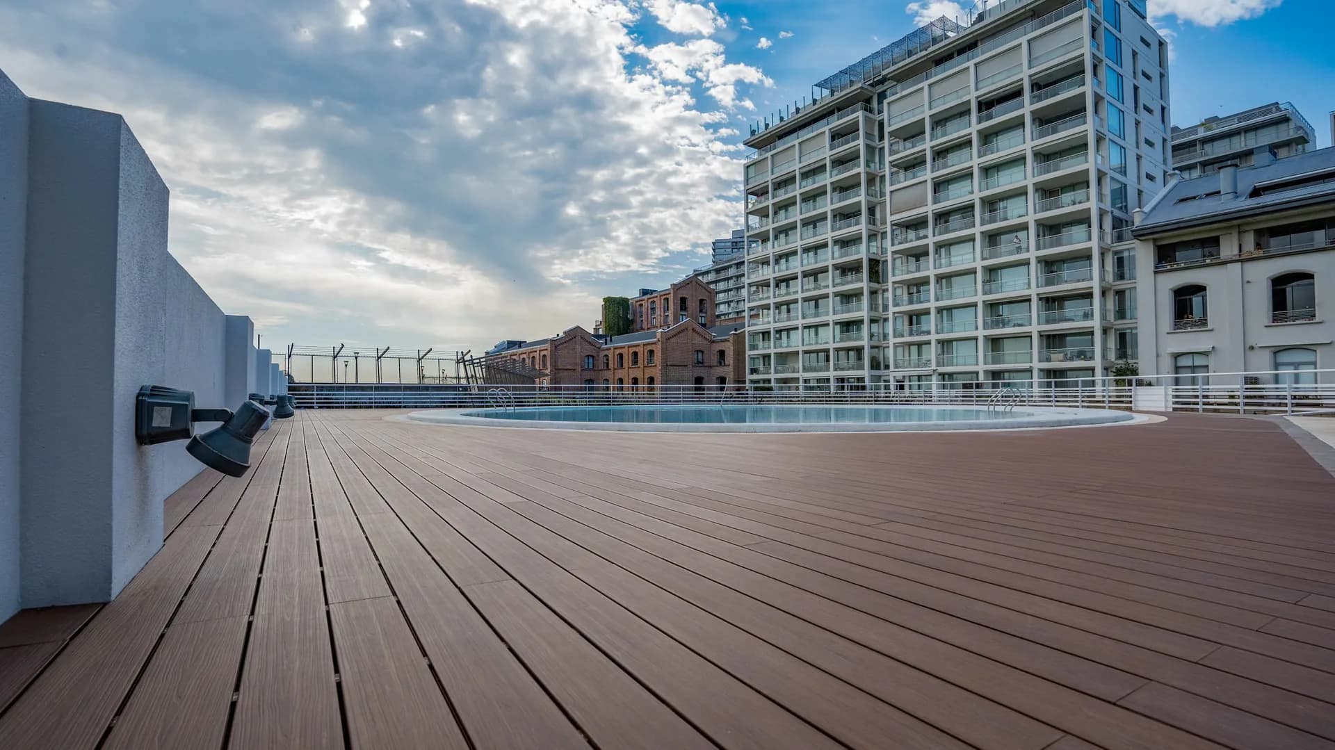 Rooftop with a pool and a wide brown WPC deck in front of apartment buildings, under a cloudy sky
