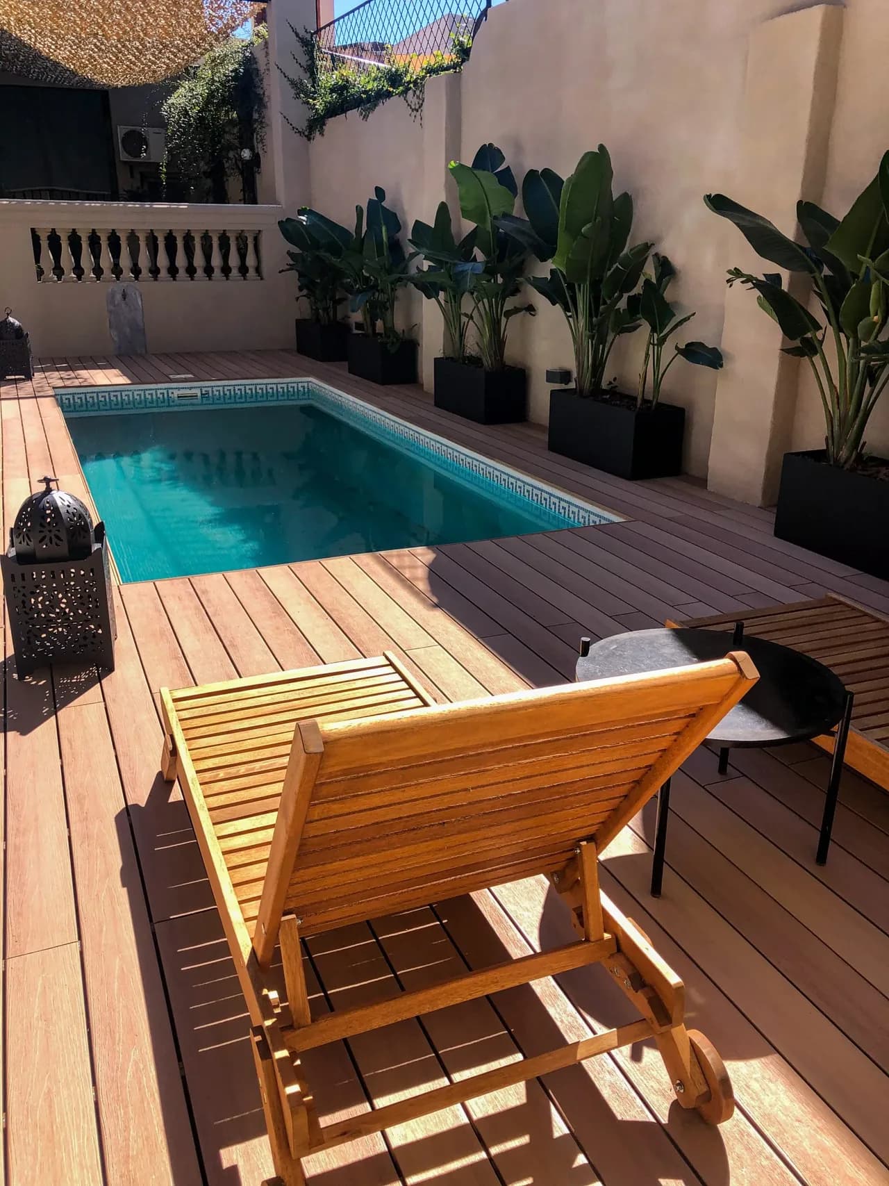 Pool in a sunny courtyard surrounded by brown WPC decking, with a wooden lounger and potted plants