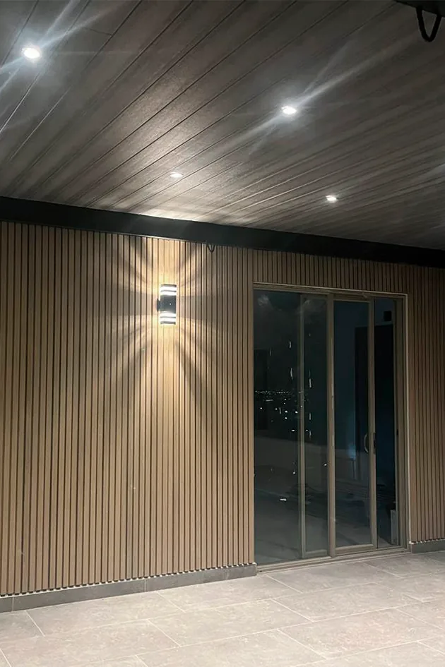 Covered patio at night with a WPC grooved panel wall, a wall light on and a glass door