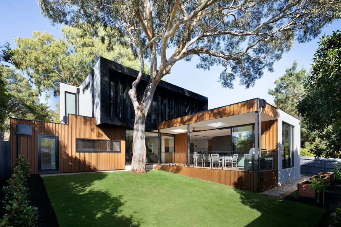 Single-story house with a wood-tone WPC grooved panel facade and an open patio, seen from the yard past a eucalyptus tree