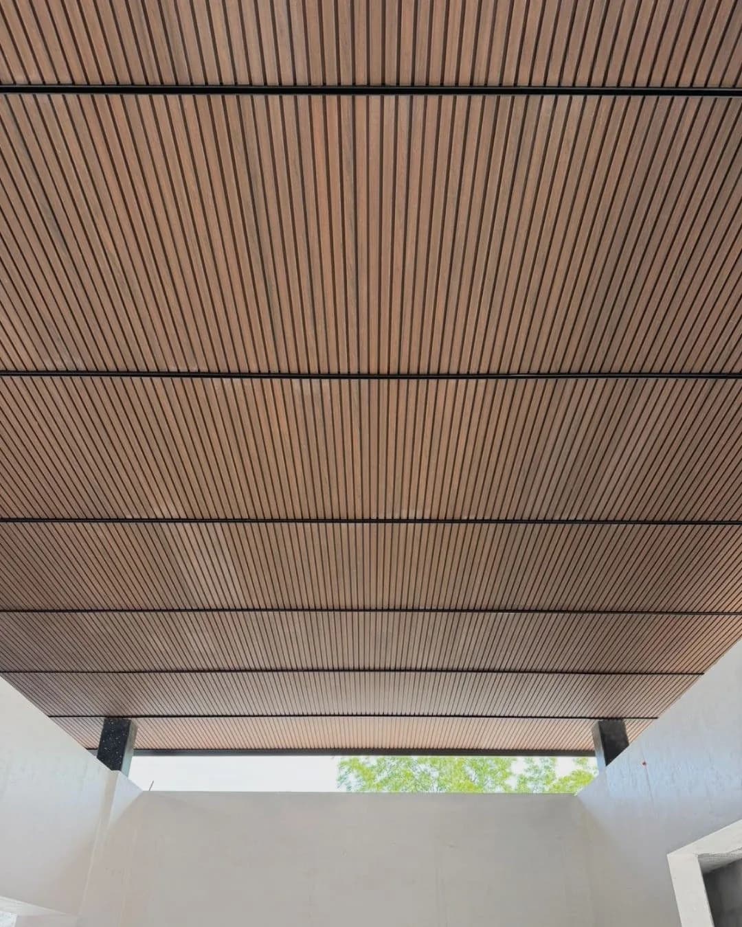 Techo exterior de un patio visto desde abajo, revestido de panel WPC ranurado tono madera entre vigas oscuras
