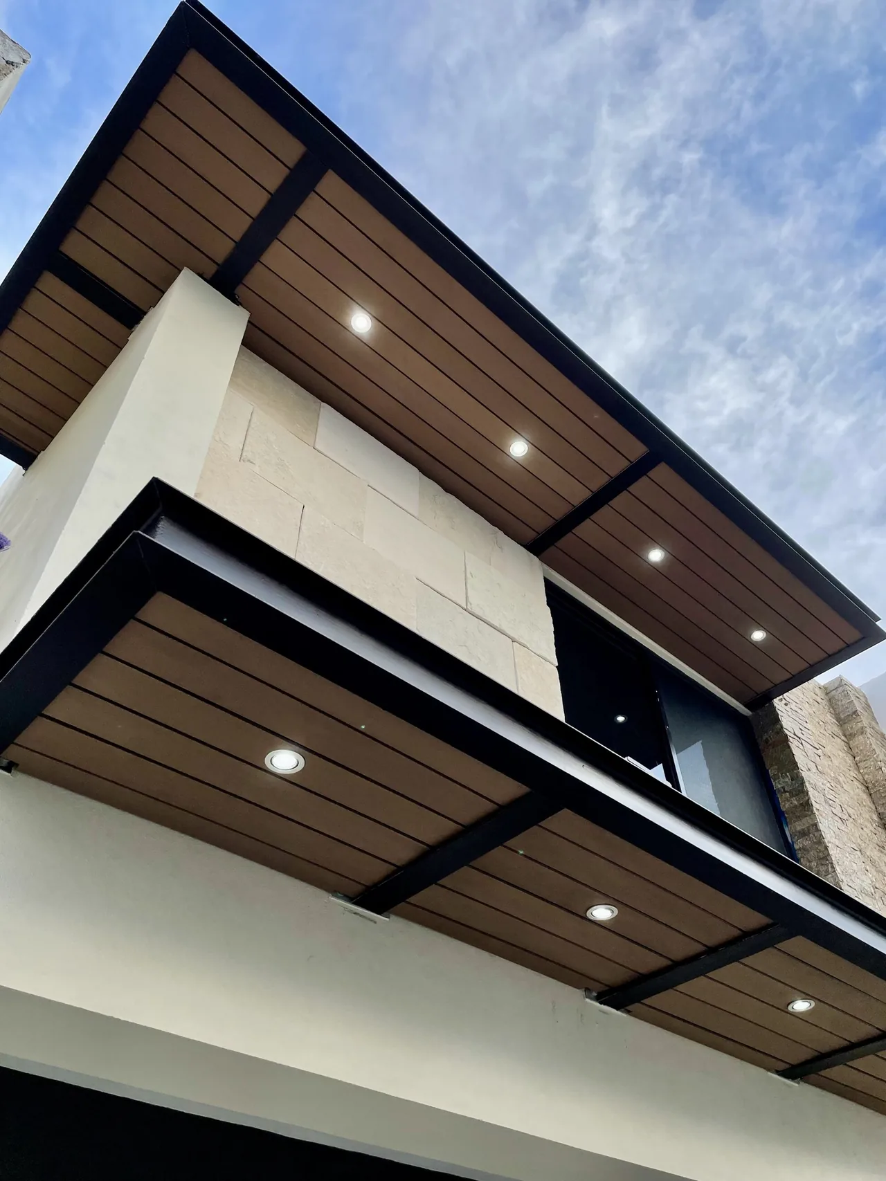 Two overhangs with wood-tone WPC soffits and recessed lights, seen from below against the sky