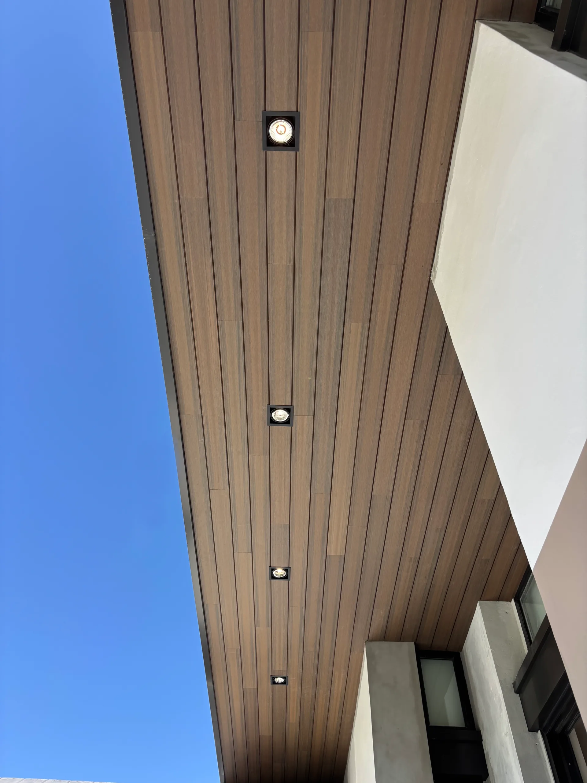 Wood-tone WPC soffit seen from below on the eave of a white facade, with recessed lights and blue sky