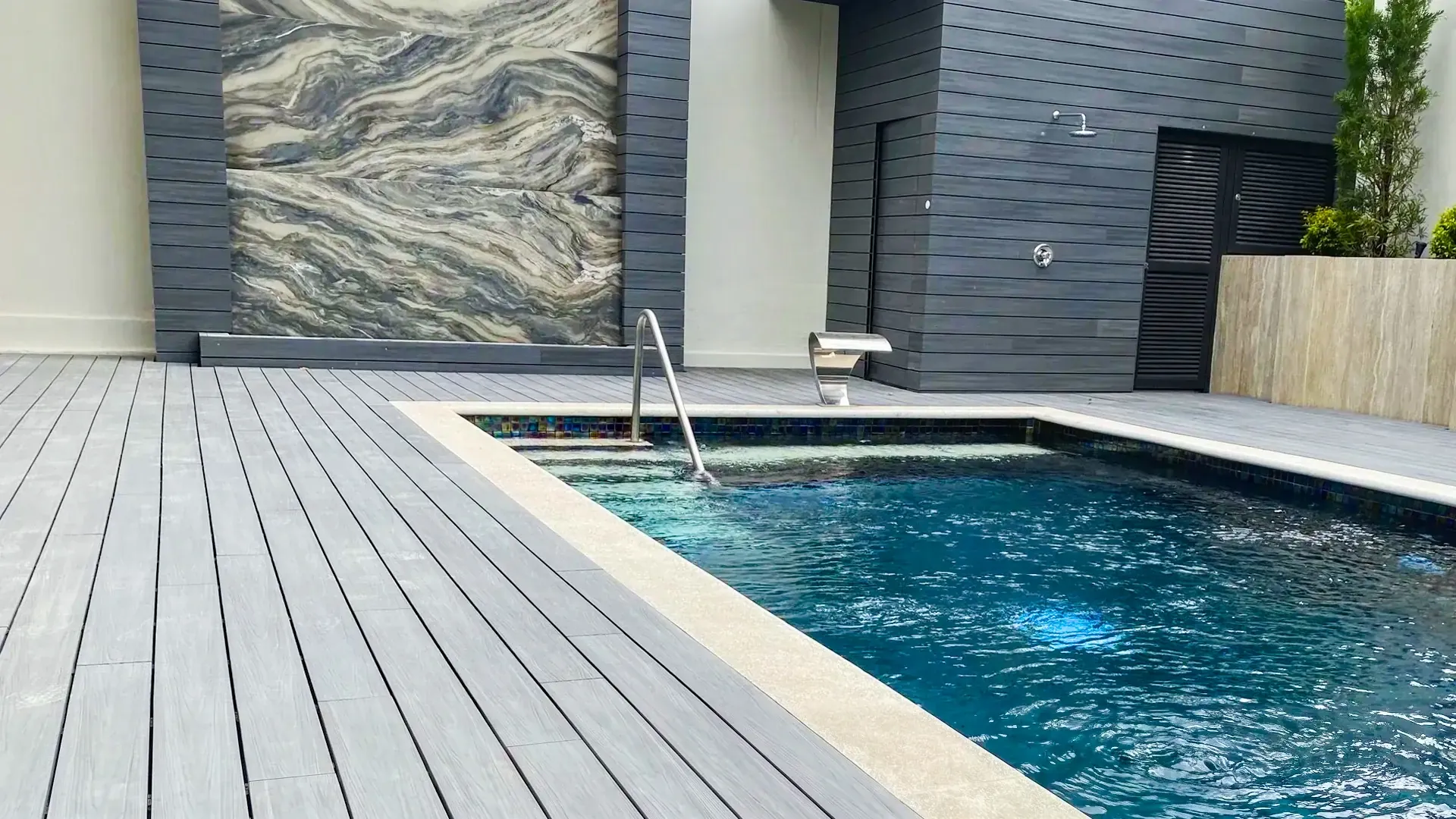 Gray WPC decking at the edge of a pool with a ladder, next to a marble wall and a gray paneled wall with an outdoor shower