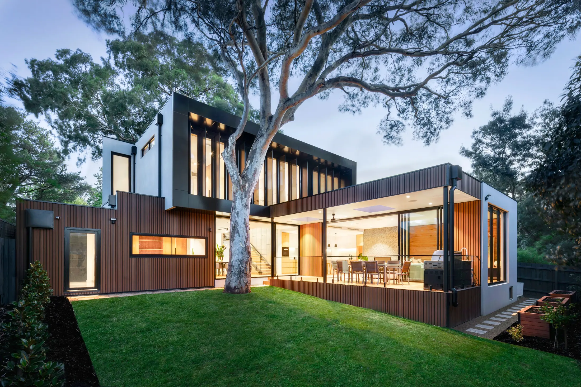Modern house at dusk with a wood-tone WPC grooved panel facade, a lit terrace and a large tree in the garden