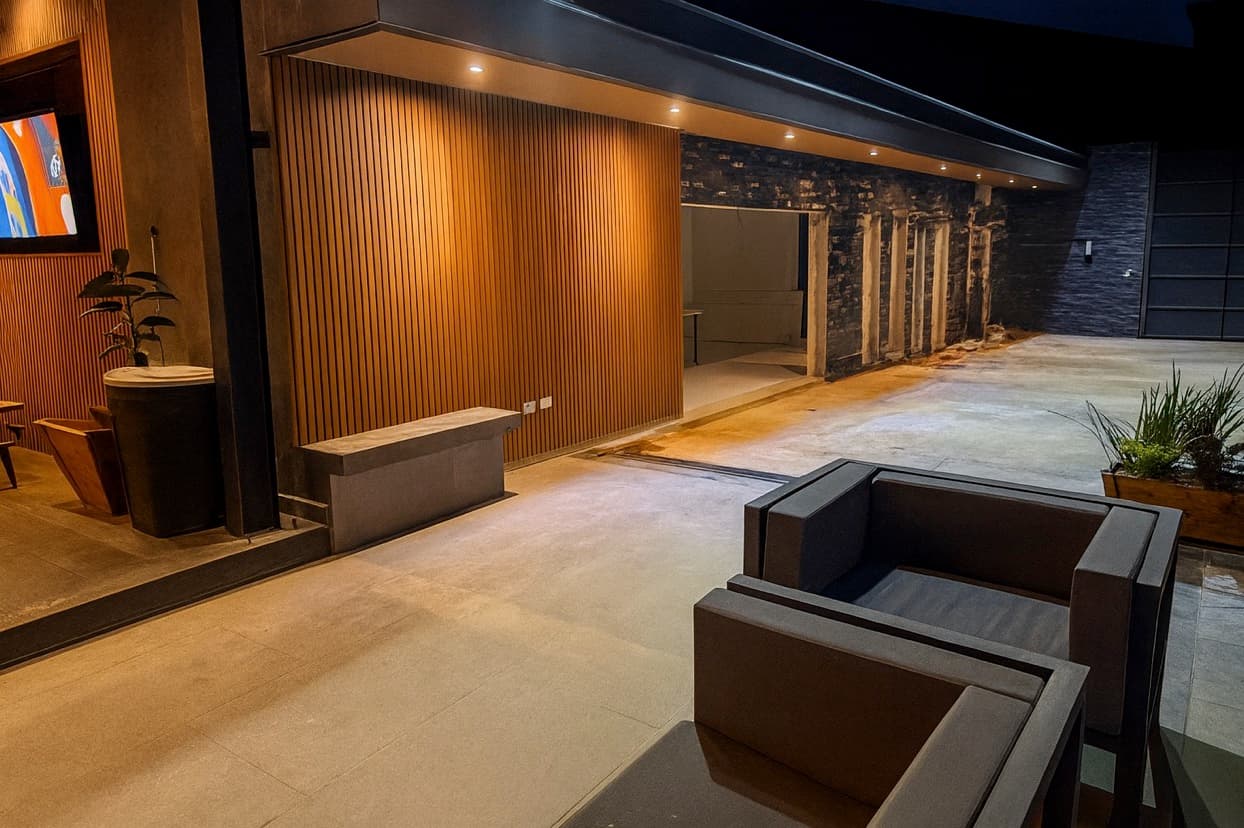 Covered terrace at night with a lit wood-tone WPC grooved panel wall under the eave, armchairs and a planter