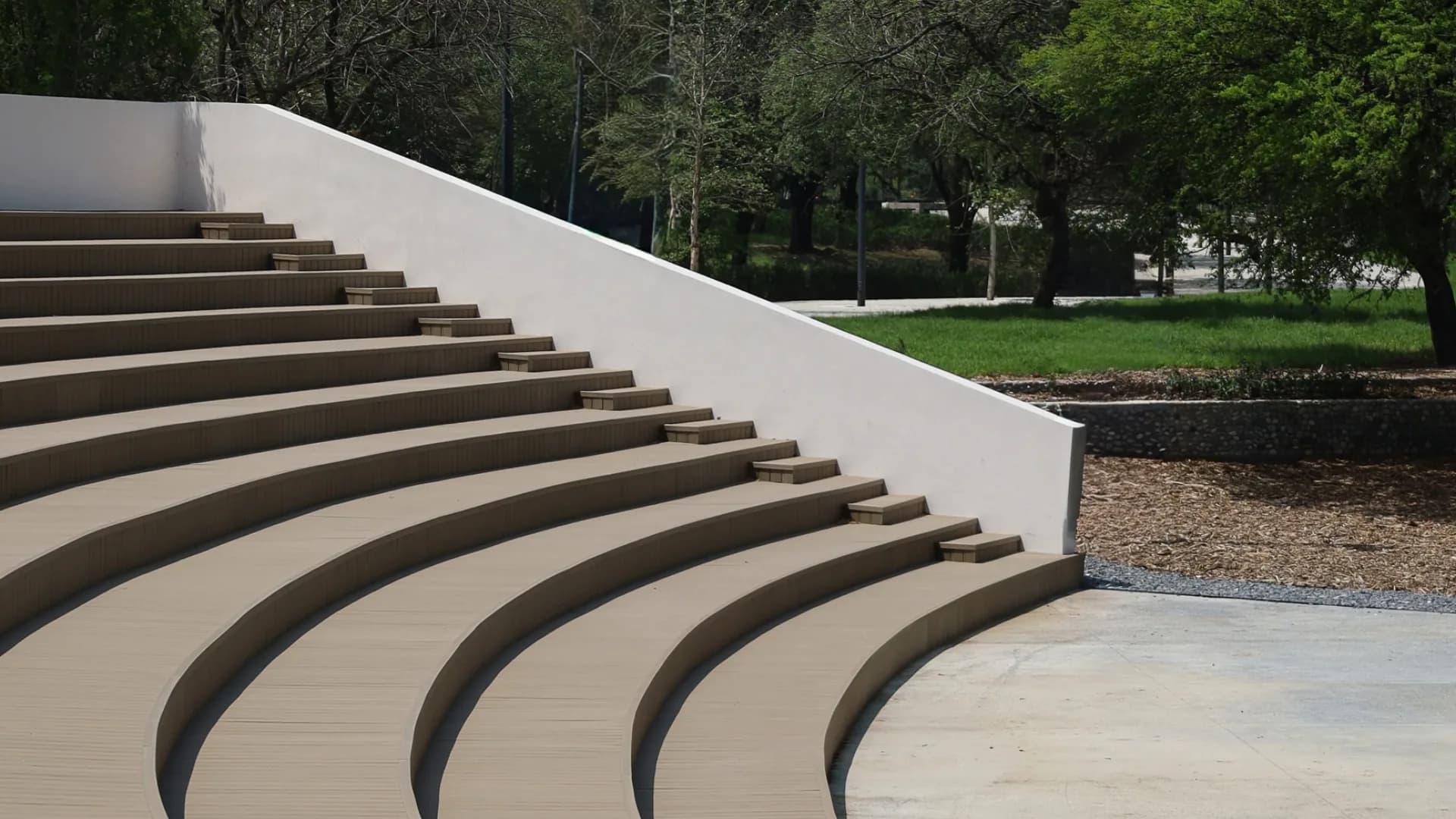 Graderío curvo de deck WPC en el Parque del Agua, junto a un muro blanco y árboles