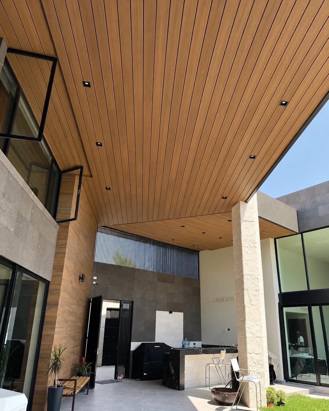 Patio cubierto de una casa con el techo revestido de Wall Cladding WPC tono madera, luces empotradas y una columna de piedra