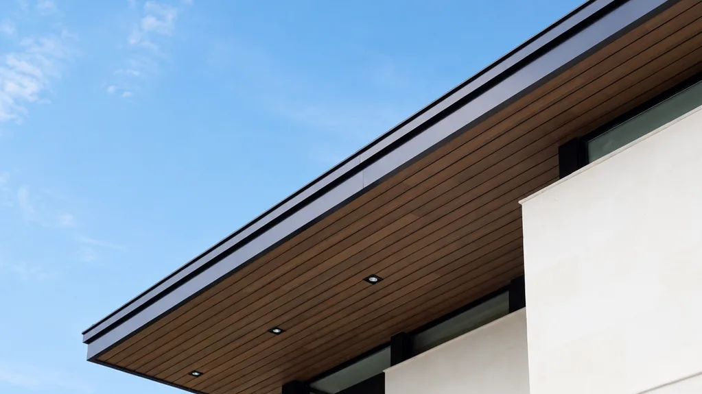 Volado de la fachada de una casa blanca revestido por debajo de Wall Cladding WPC tono madera con luces empotradas, contra cielo azul