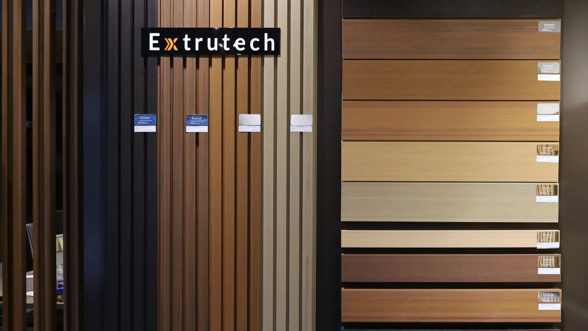 Exhibidor de Extrutech en tienda: tiras verticales de lambrín WPC en varios colores bajo el logo y, a la derecha, tablas WPC en ocho tonos, cada una con su etiqueta