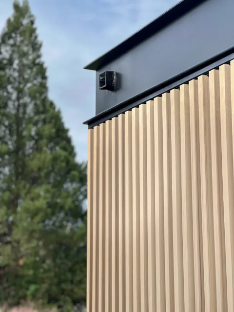 Facade corner with vertical sand-tone WPC grooved panels next to a gray section, with pine trees behind