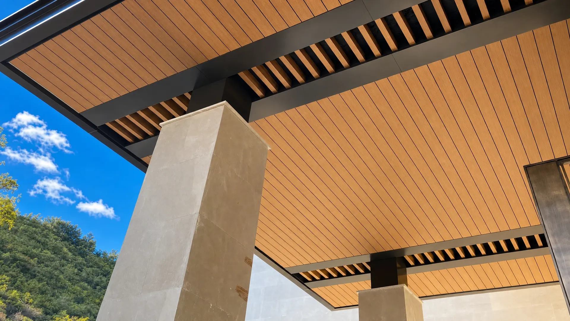 Plafón WPC tono madera con vigas y celosía sobre columnas de piedra clara, con cielo azul y un cerro con vegetación