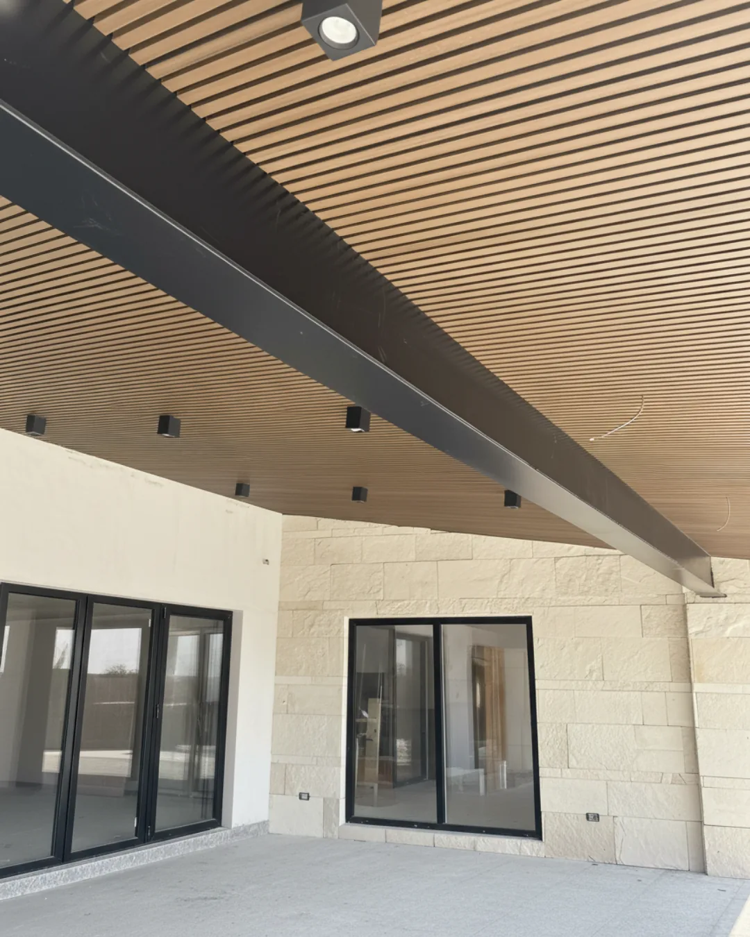 Terraza de una casa con techo de Wall Cladding (Plafón) tono madera y luces empotradas, sobre un muro de piedra clara y ventanales
